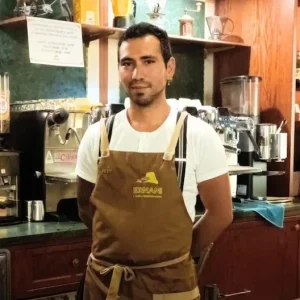 Foto di Cristian, un barista della Torrefazione Ernani a Milano con la divisa estiva (grembiule Ernani e maglietta bianca mezze maniche) dietro al bancone del locale.