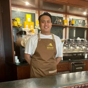 Foto di Mark, un barista della Torrefazione Ernani a Milano con la divisa estiva (grembiule Ernani e maglietta bianca mezze maniche) dietro al bancone del locale.